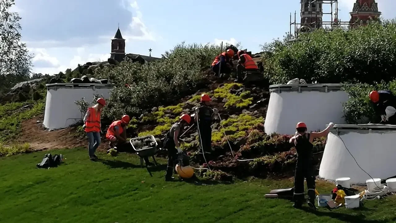 Рабочие в касках и жилетах сажают растения на склоне, рядом бетонные кольца и тачка